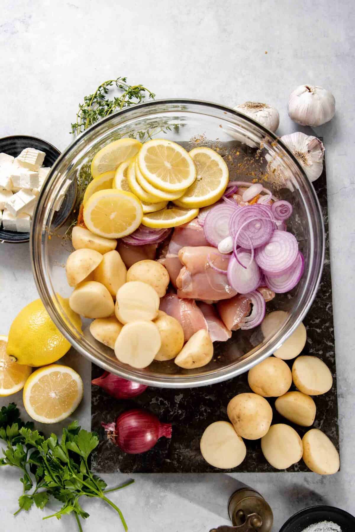 A glass bowl filled with raw chicken, halved potatoes, lemon slices, and sliced red onions. Surrounding the bowl are whole potatoes, garlic, lemons, red onions, cheese cubes, herbs, and greenery.