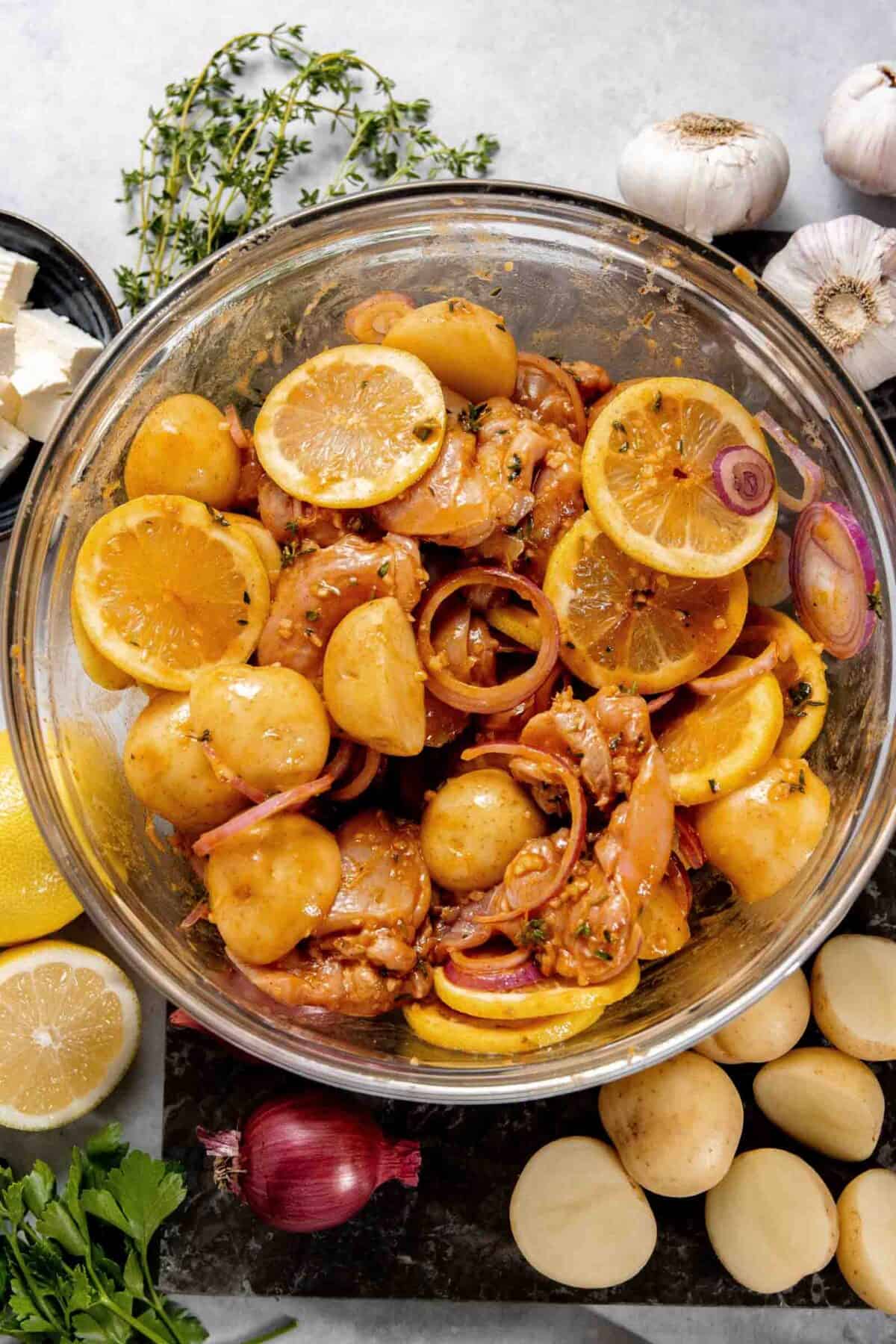 A glass bowl filled with seasoned chicken pieces, potato halves, and lemon slices, placed on a countertop with garlic, onion, lemon, butter, and herbs scattered around.