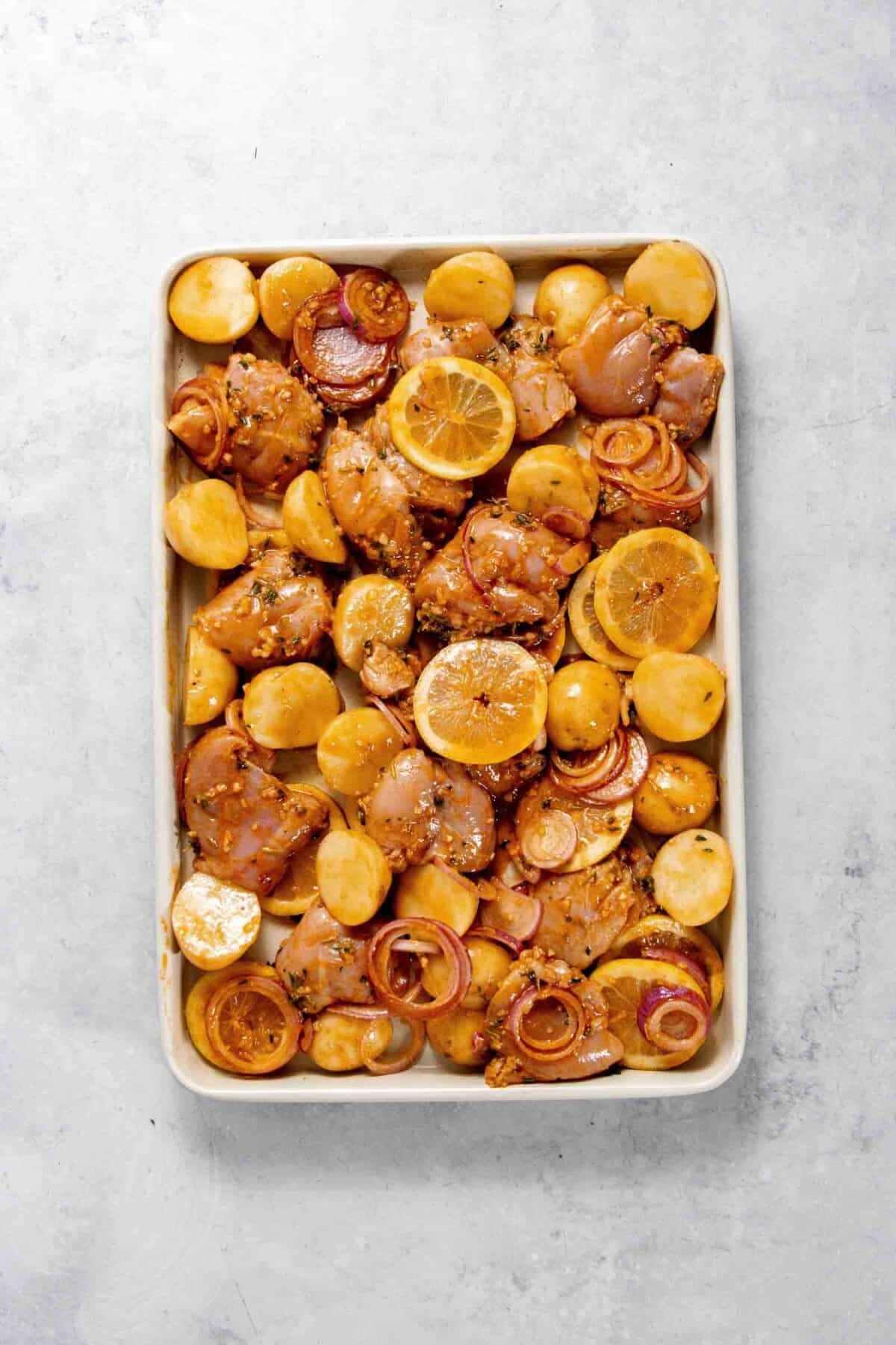 A baking tray containing seasoned chicken pieces, sliced baby potatoes, red onions, and lemon slices arranged ready for roasting.