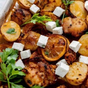 Close-up of a dish featuring roasted chicken, potatoes, lemon slices, and cubes of feta cheese, garnished with fresh parsley.