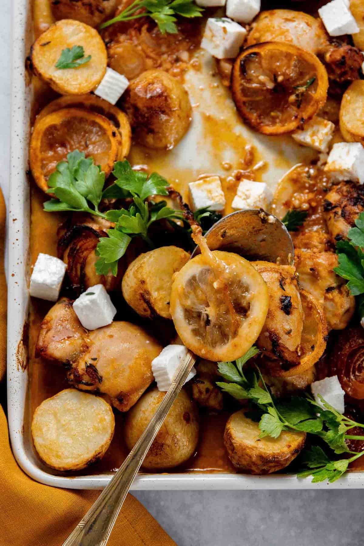 A roasted dish featuring slices of lemon, potatoes, chicken pieces, and cubes of white cheese garnished with parsley on a baking tray.