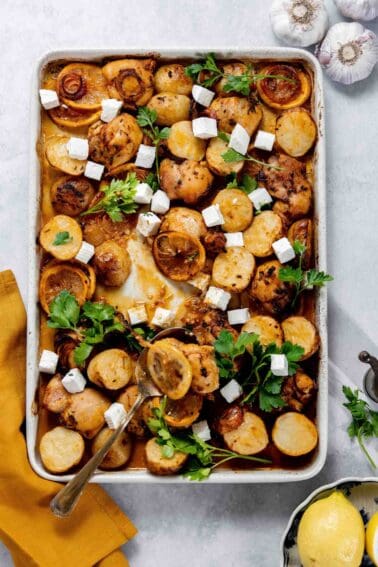 A tray with roasted potatoes, onions, and herbs, topped with chunks of cheese. A spoon rests inside the tray. Garlic bulbs and a lemon are next to the tray.