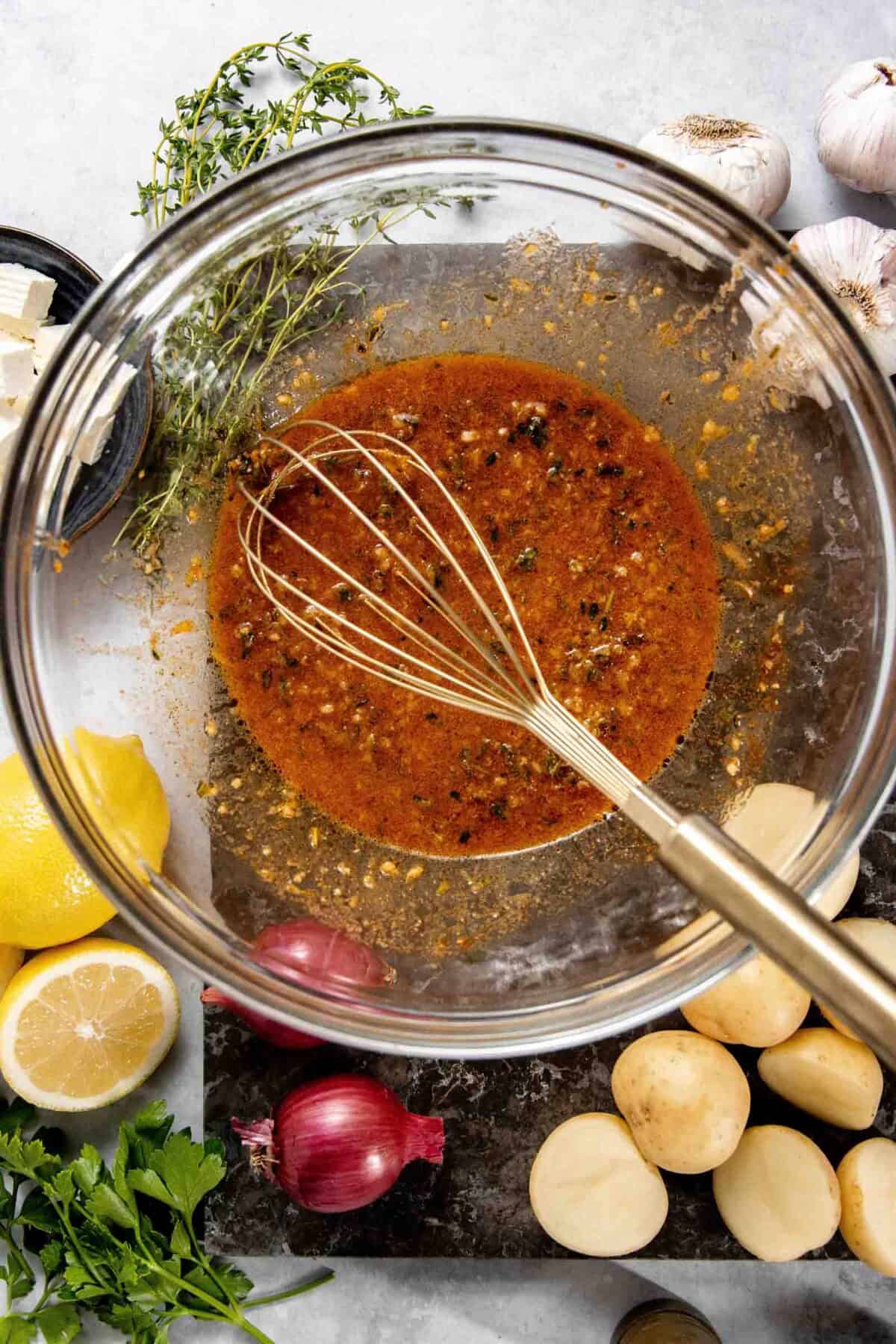 A glass bowl filled with a seasoned marinade, being whisked, surrounded by herbs, garlic, onions, lemons, and potatoes.