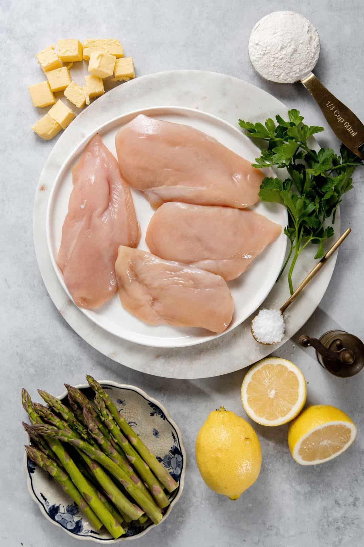 Raw chicken breasts on a plate with ingredients including butter, flour, parsley, salt, asparagus, and lemons arranged around on a marble surface.