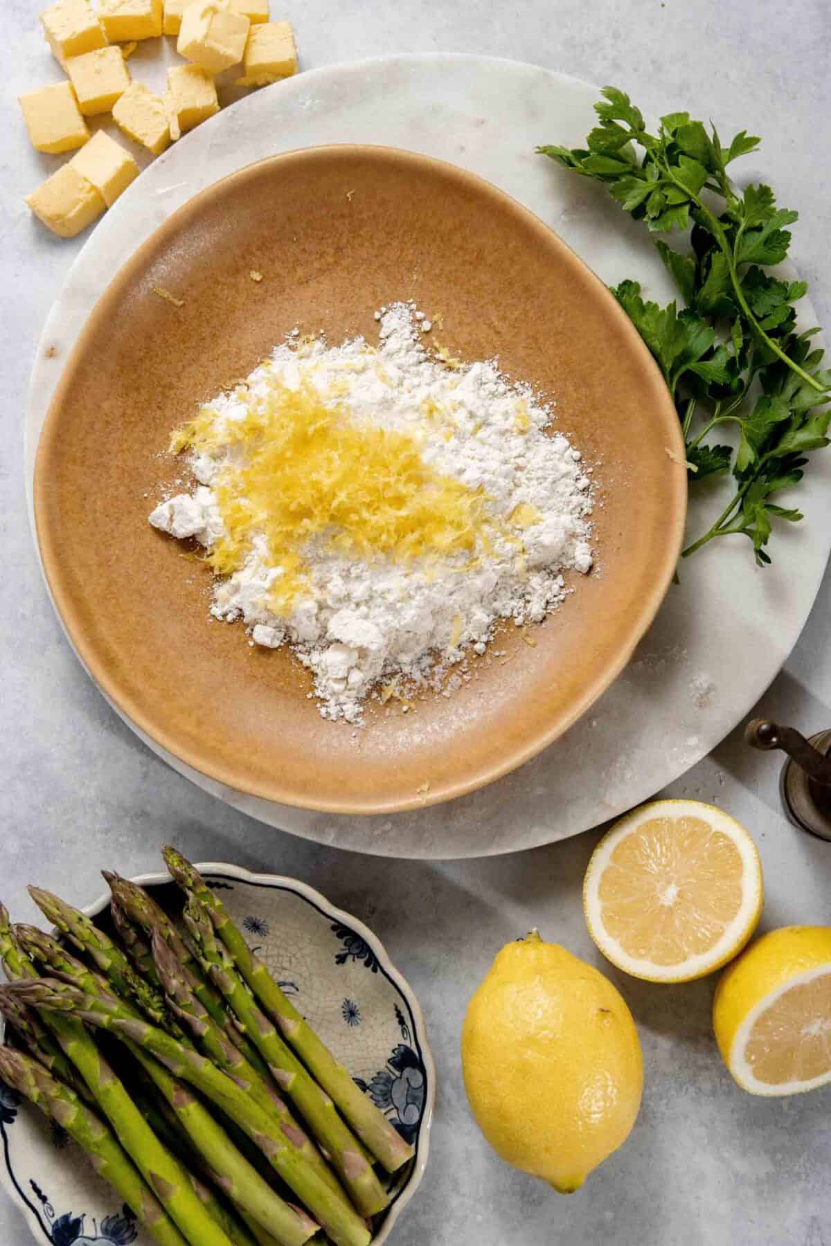 A large bowl containing flour and lemon zest sits on a marble surface, surrounded by asparagus, lemon halves, parsley, and small cubes of butter.