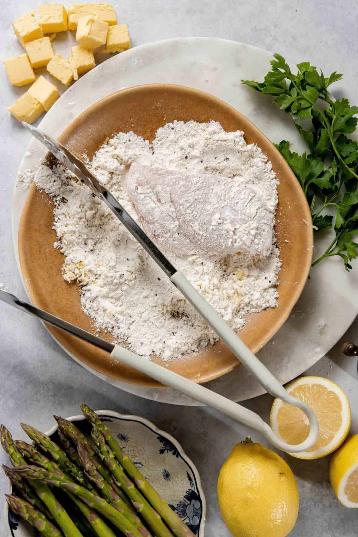 A piece of chicken is being breaded with flour on a brown plate, surrounded by lemon halves, cubes of butter, parsley, asparagus spears, and kitchen tongs on a white surface.