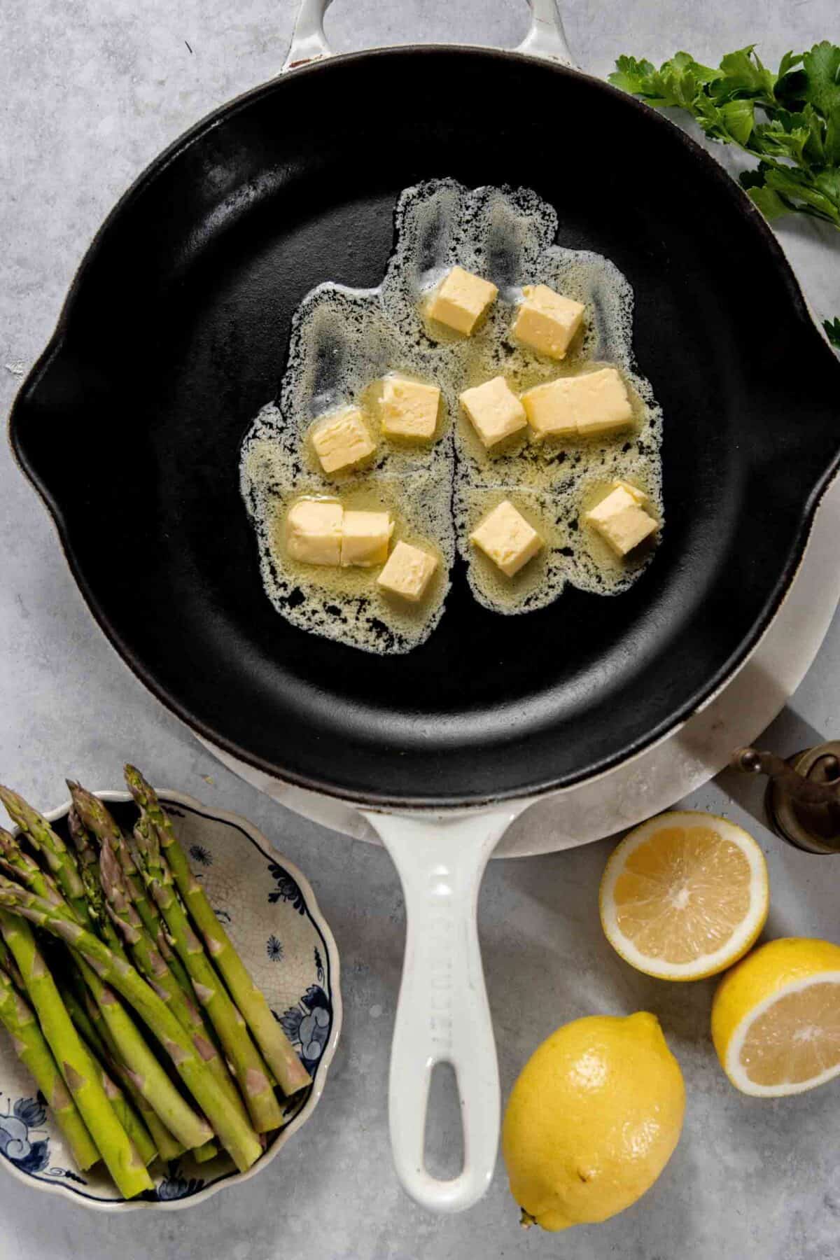 A cast iron skillet with melting butter cubes, surrounded by asparagus, two lemon halves, a whole lemon, and green herbs.