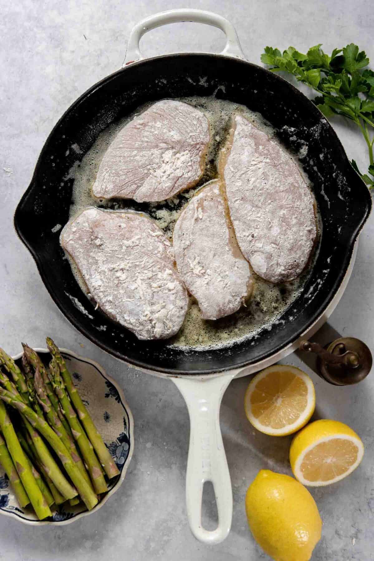 Raw, flour-coated chicken pieces in a frying pan with melted butter. A small bowl of asparagus, two lemon halves, a whole lemon, and parsley are beside the pan on a marble surface.