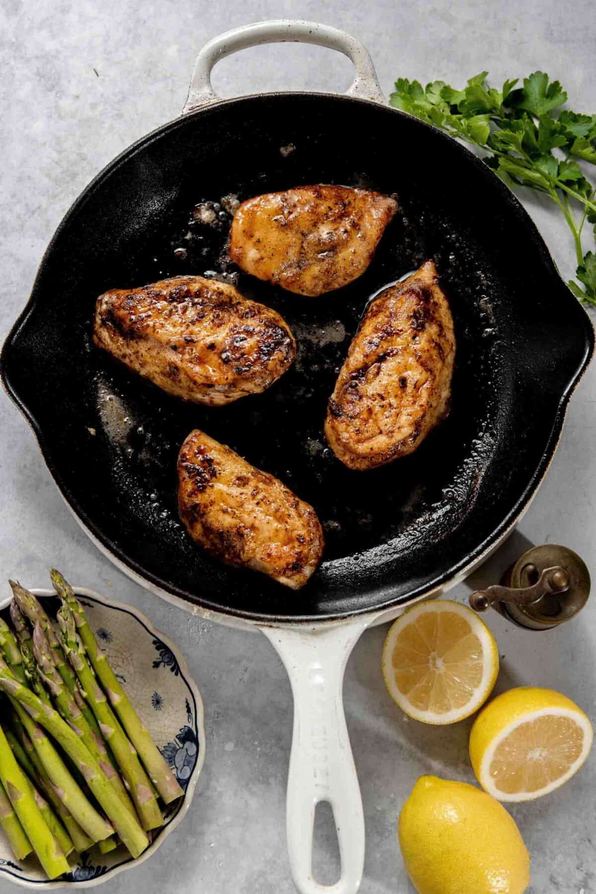 Four cooked chicken breasts in a white cast iron skillet, with a plate of asparagus, two lemons, parsley, and a pepper grinder arranged nearby on a light surface.