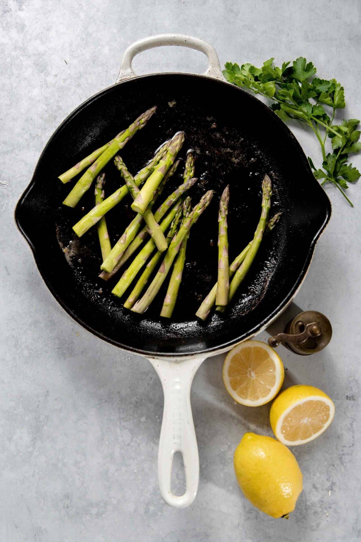 A white cast-iron skillet with asparagus spears is placed on a grey surface alongside halved lemons, a salt shaker, and a sprig of parsley.