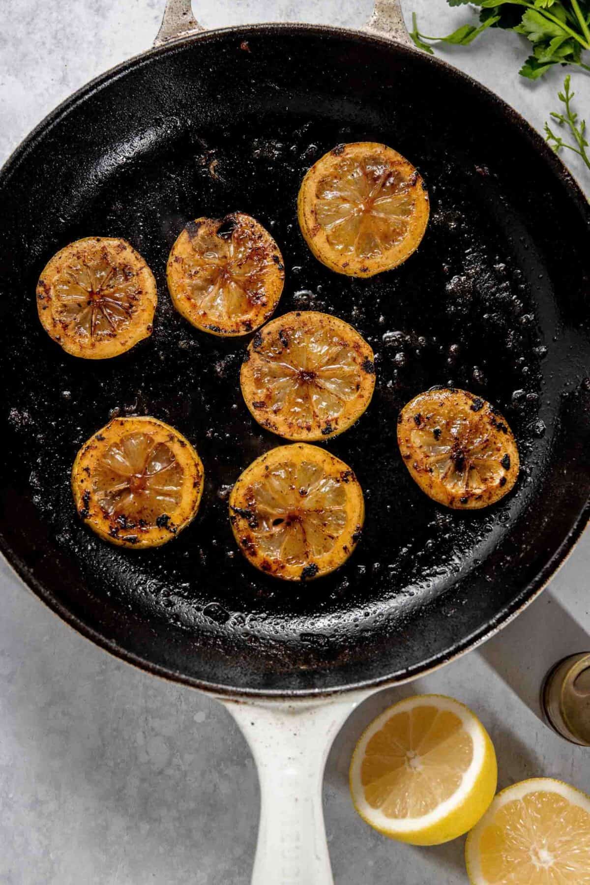 A cast iron skillet with seven slices of seared lemon. Two halves of a fresh lemon and sprigs of herbs are placed near the skillet on a light-colored surface.