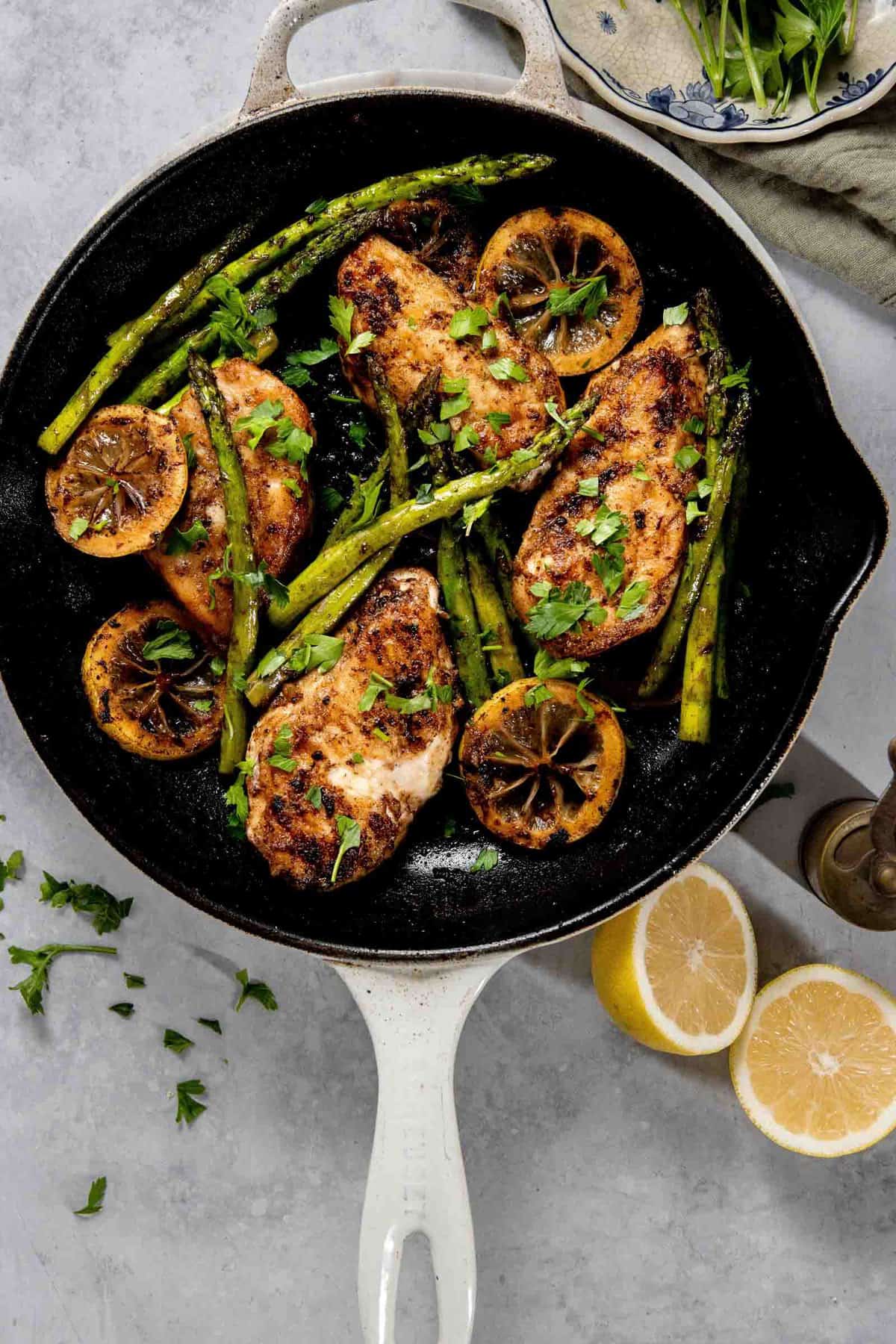 A skillet with cooked chicken breasts, asparagus, and lemon slices garnished with fresh herbs. A sliced lemon and a dish with greens are visible beside the skillet on a light-colored surface.