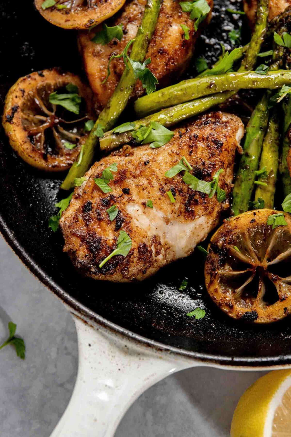 Seared chicken breast with charred lemon slices and asparagus spears in a cast iron skillet, garnished with chopped parsley.