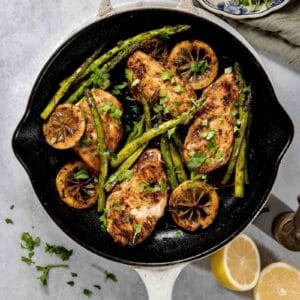 A skillet containing cooked chicken breasts, lemon slices, and asparagus, garnished with fresh herbs. Lemons and a bowl with leafy greens are positioned nearby.