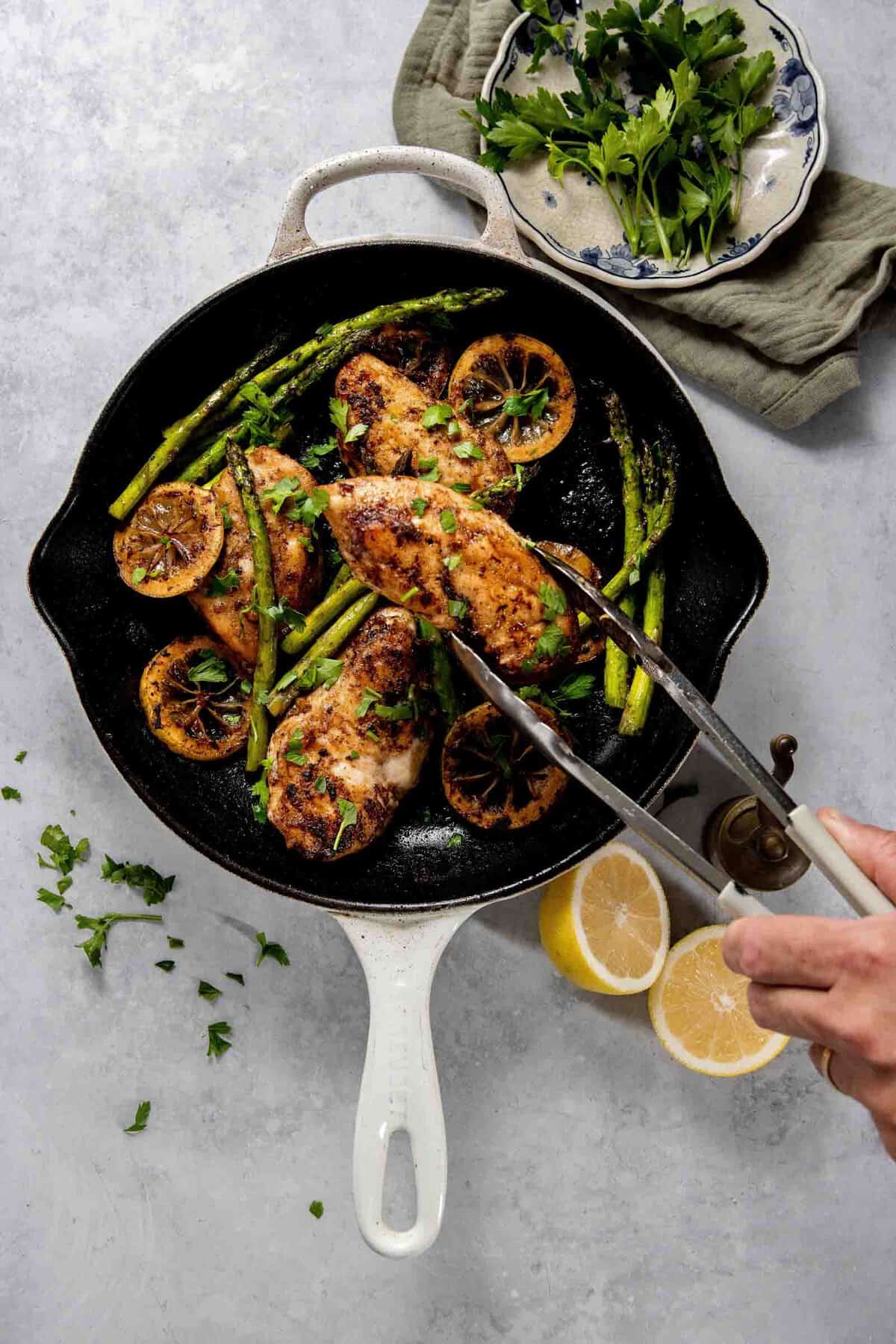 A skillet with chicken breasts topped with herbs, surrounded by asparagus and seared lemon slices. Beside the skillet are a plate with parsley, a cloth napkin, and halved lemons.