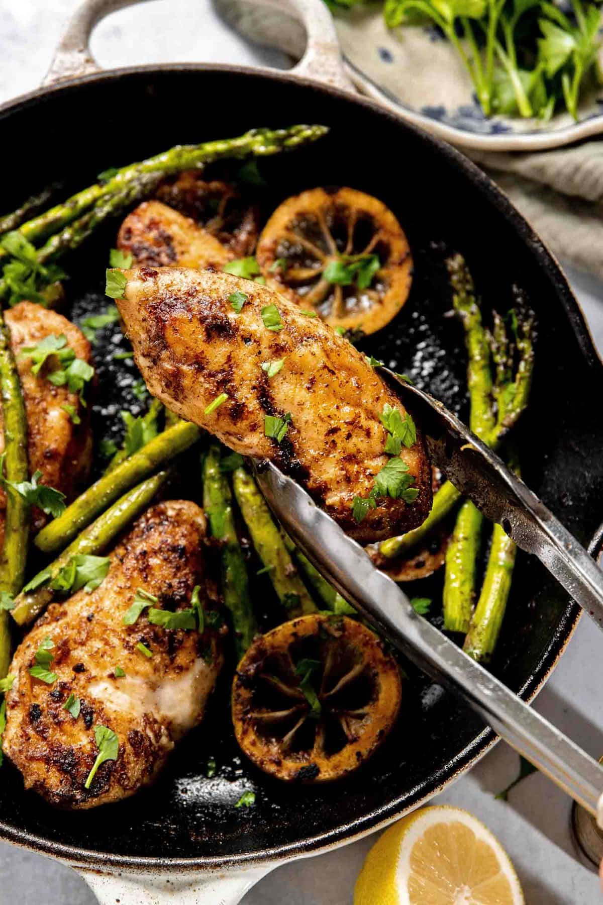 A cast iron skillet containing seasoned chicken breasts, asparagus, lemon slices, and garnished with fresh herbs, with a set of metal tongs holding one piece of chicken.