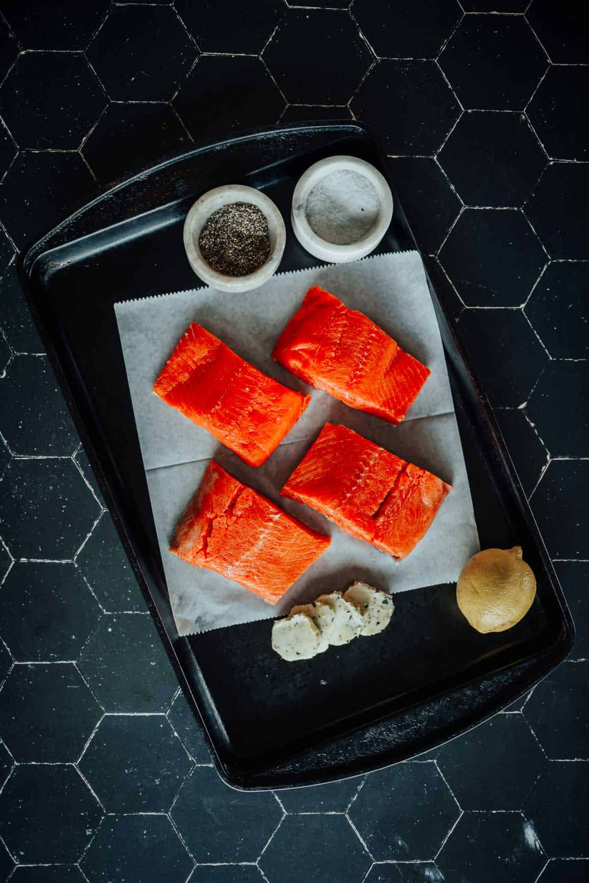 Four raw salmon fillets on parchment paper in a baking tray, surrounded by bowls of salt and pepper, a lemon, and small dollops of butter on a dark background.