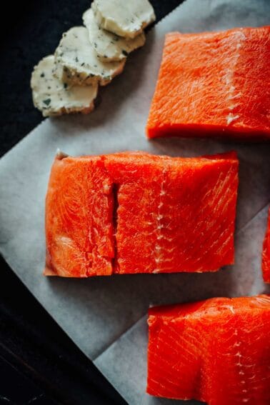 Close-up of raw salmon fillets on white parchment paper with slices of herb butter on the side.