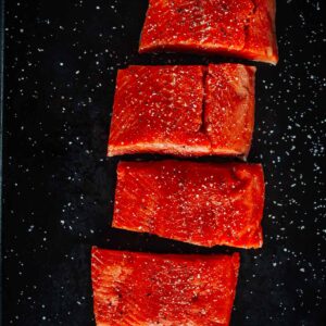Four raw salmon fillets, evenly spaced, placed on a black surface with coarse salt scattered around.