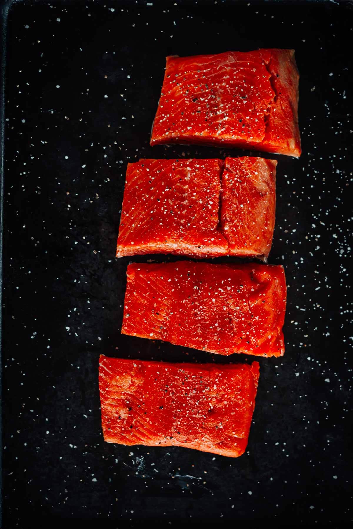 Four raw salmon fillets, evenly spaced, placed on a black surface with coarse salt scattered around.