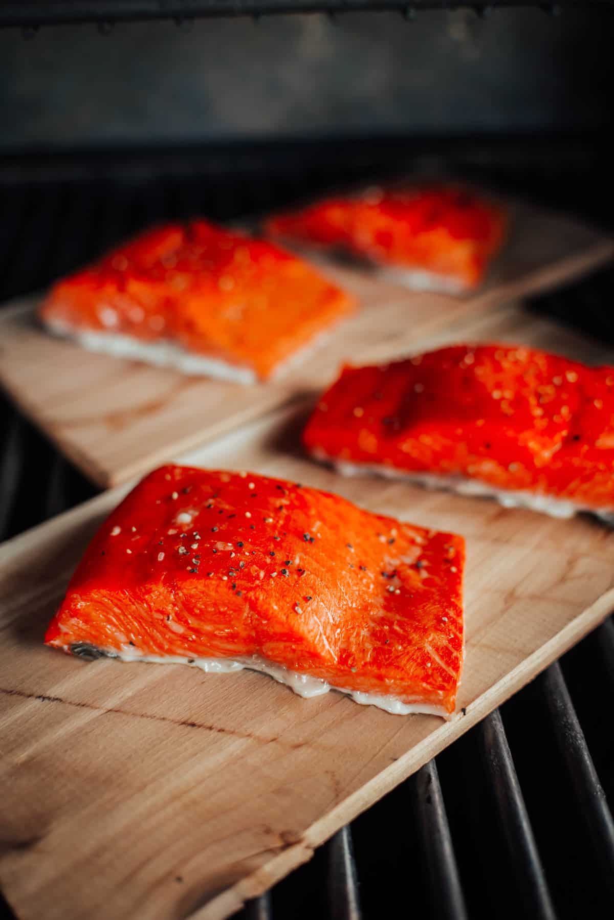 Several salmon fillets seasoned with spices are placed on wooden planks cooking on a grill.