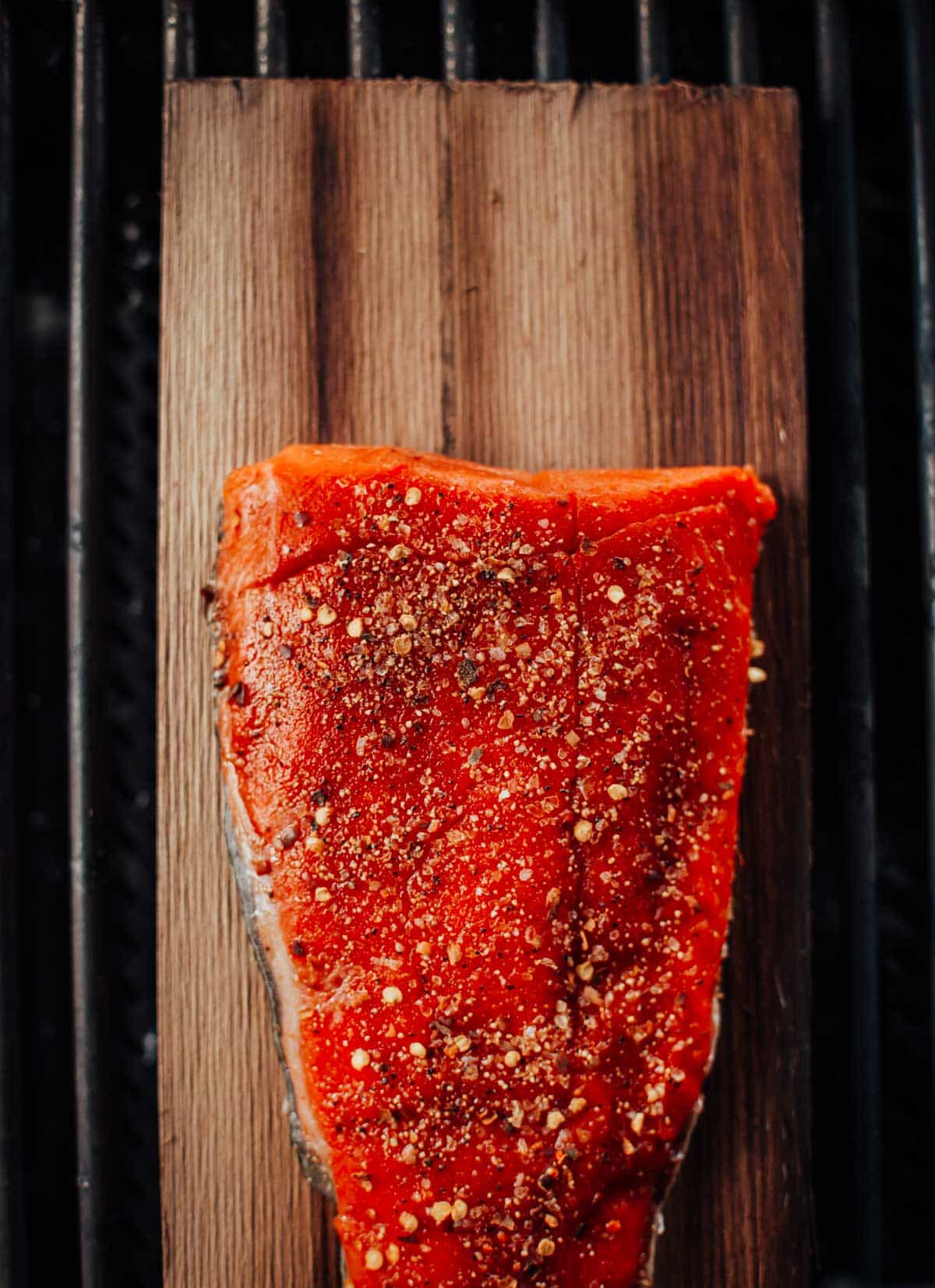 A seasoned, raw salmon fillet on a wooden plank, positioned on a grill.