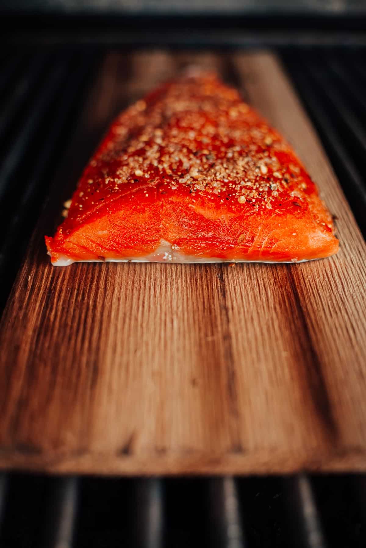 A seasoned salmon fillet is placed on a wooden plank, set on a grill, ready to be cooked.