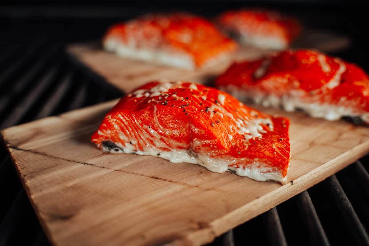 Four pieces of seasoned salmon fillets are cooking on wooden planks on a grill.