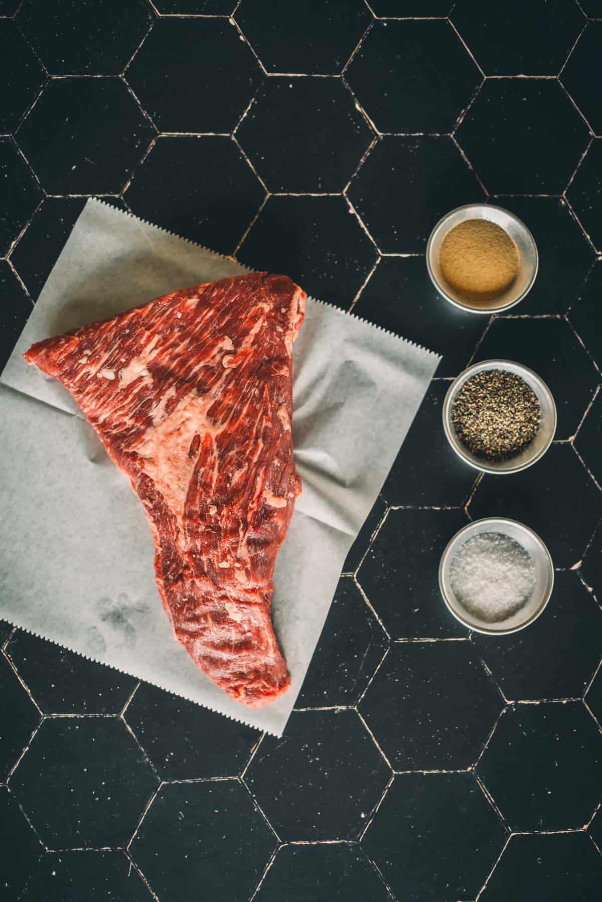 A raw tri-tip on parchment paper is positioned on a black tile surface, with small bowls containing spices placed beside it.