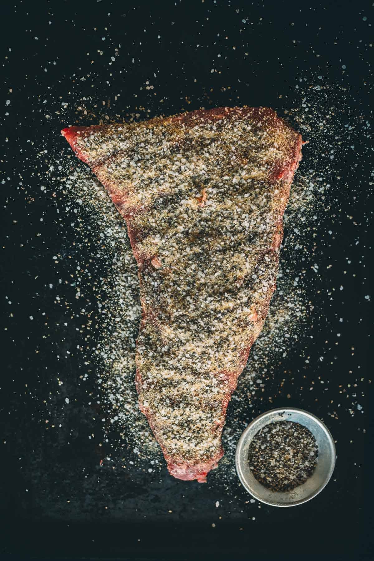 A tri-tip seasoned with spices lies on a dark surface next to a small bowl containing additional seasoning.