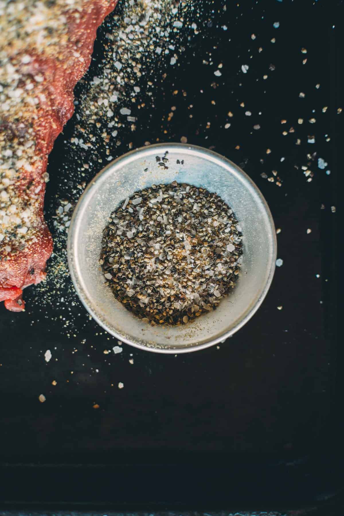 A small metal bowl filled with ground black pepper, salt and granulated garlic sits on a black surface next to a piece of raw, seasoned meat.