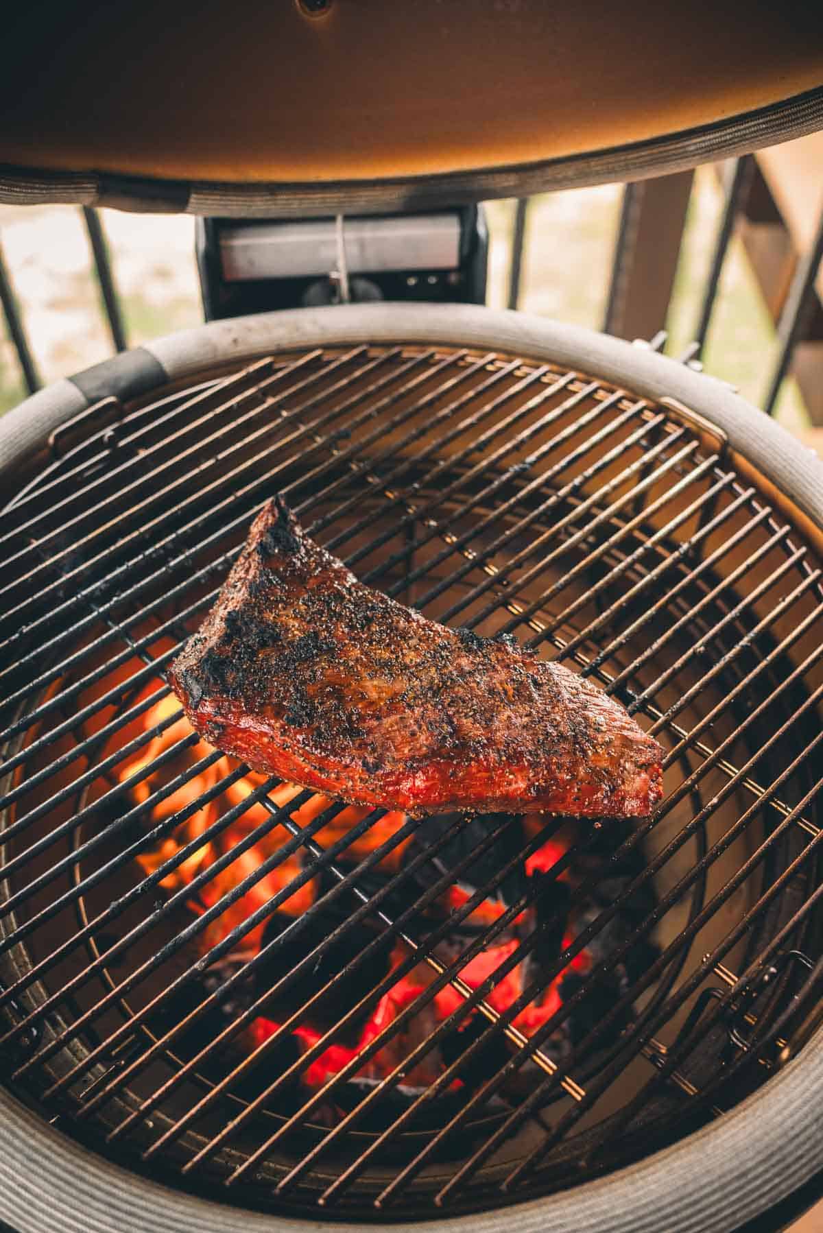 A tri-tip is being grilled on an outdoor barbecue grill, with visible flames and hot coals beneath the grill grates.