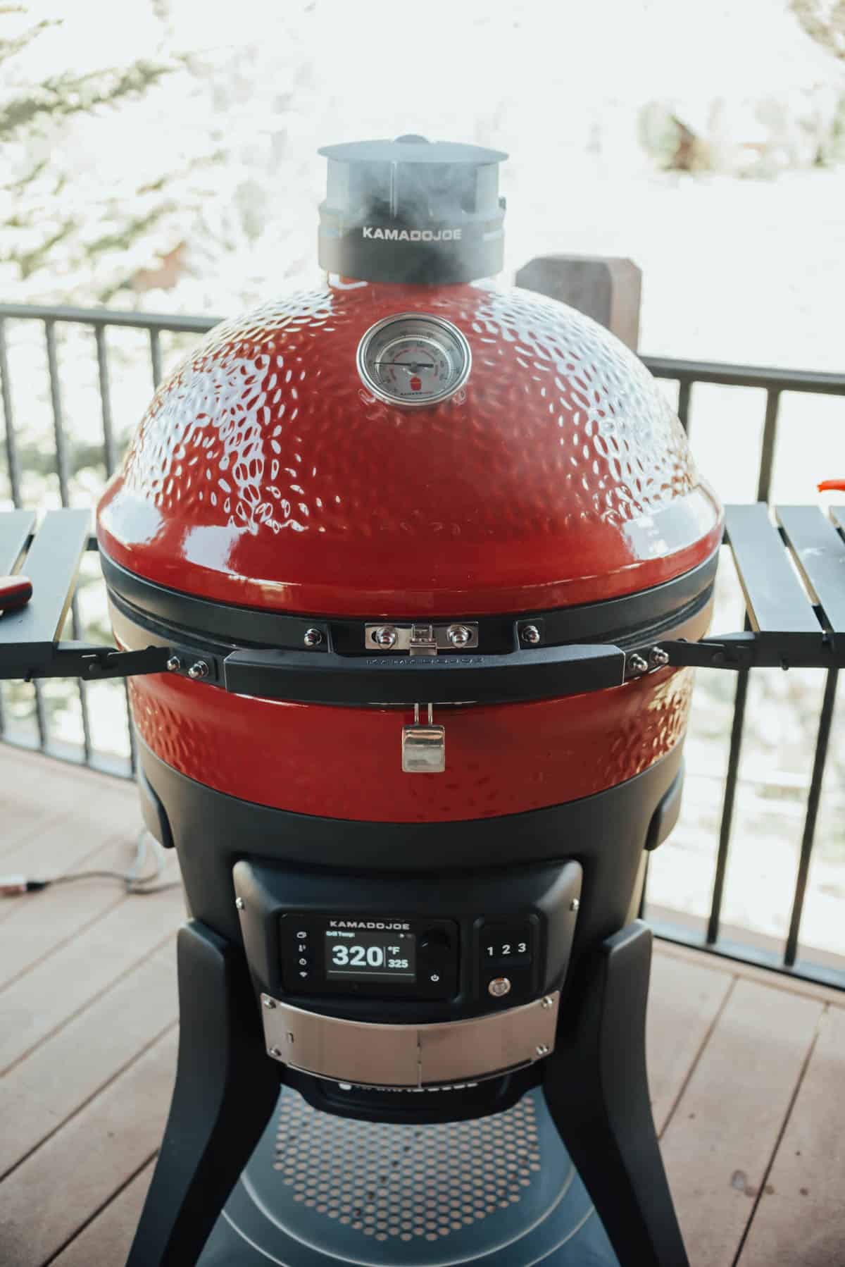 A red Kamado Joe ceramic grill with a temperature gauge on top and a digital control panel displaying 320°F, placed on an outdoor deck.