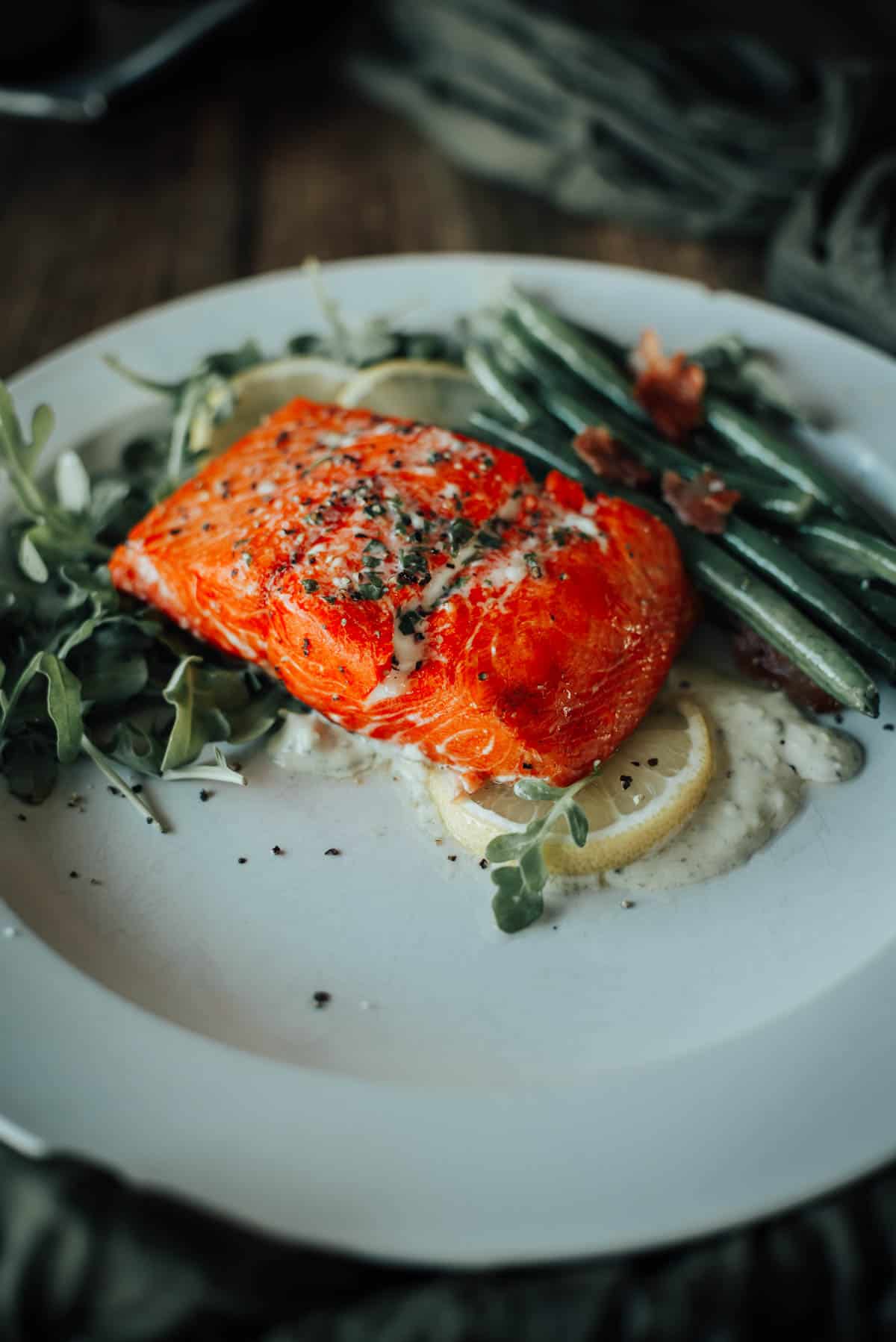 A plated smoked salmon fillet garnished with herbs, served with green beans, sliced lemon, and a dollop of sauce on a white plate.