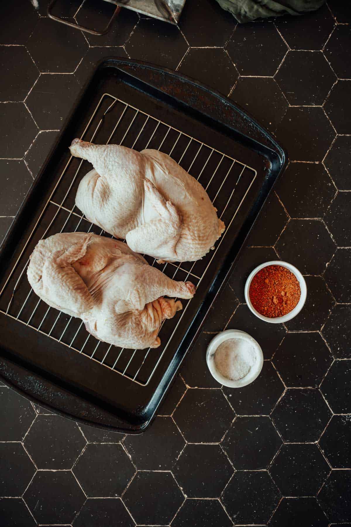 Two raw whole chickens halves on a wire rack in a baking tray with bowls of seasoning and salt on a black surface.