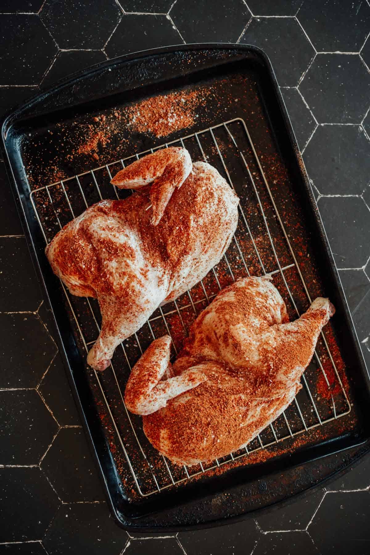 Two raw chicken halves covered in seasoning sit on a wire rack placed on a black baking sheet.