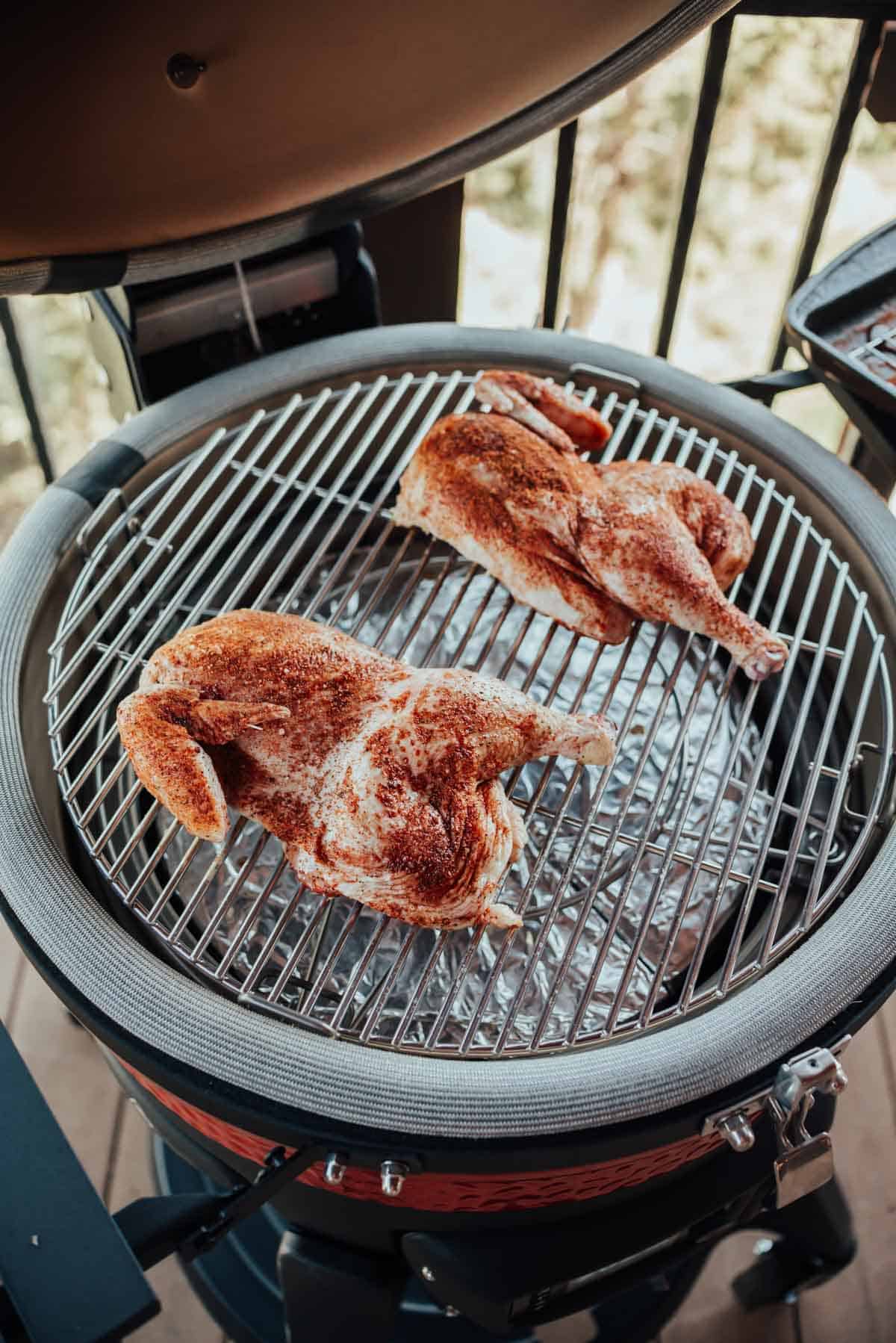 Two seasoned chicken halves being smoked on a ceramic barbecue grill.