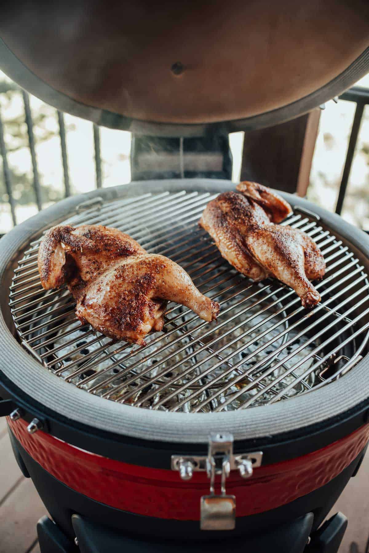 Two seasoned chicken halves are being cooked on a red ceramic charcoal grill with smoke rising from them.