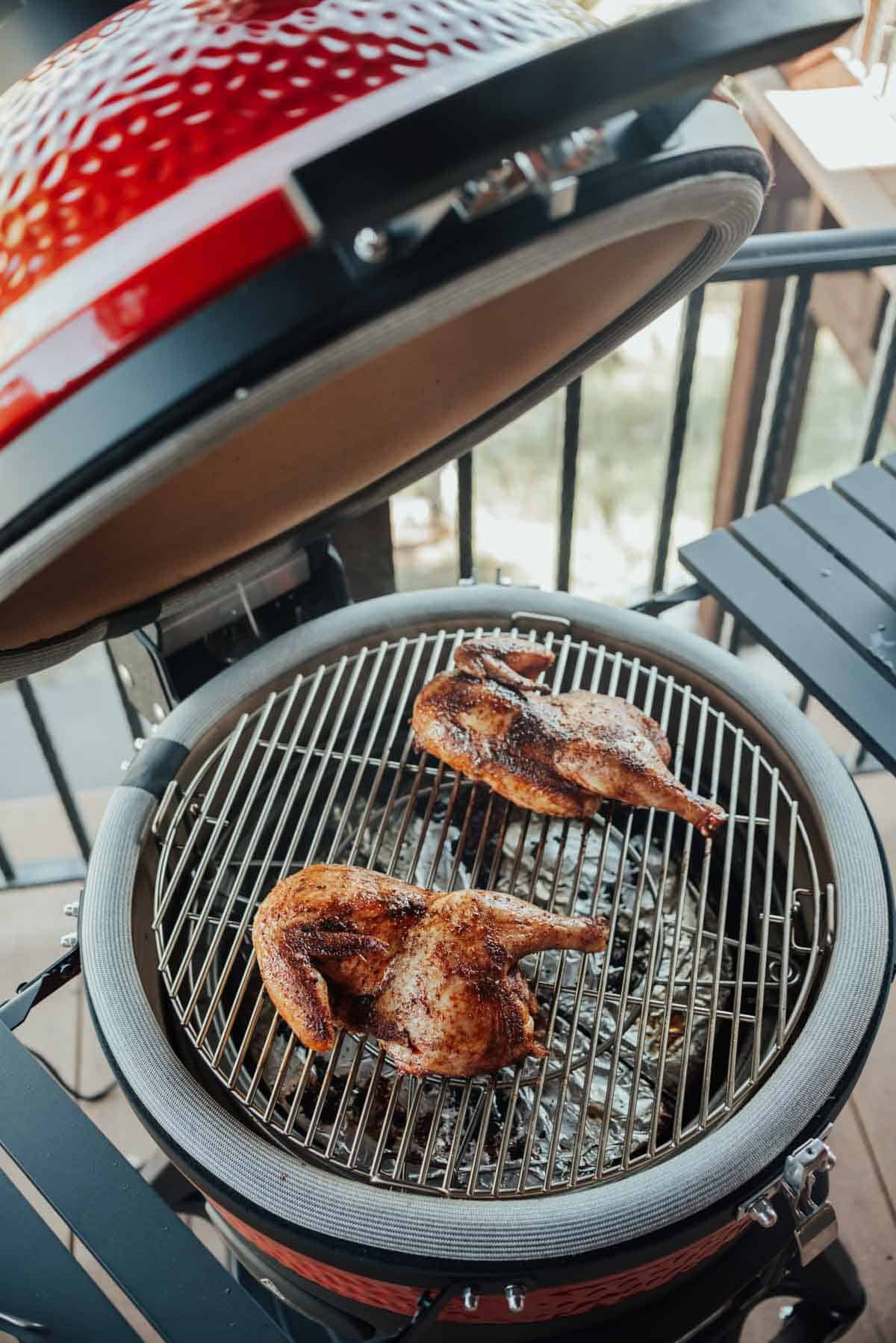 A partially open red kamado grill with two seasoned and grilled chicken halves on the grate on an outdoor patio.