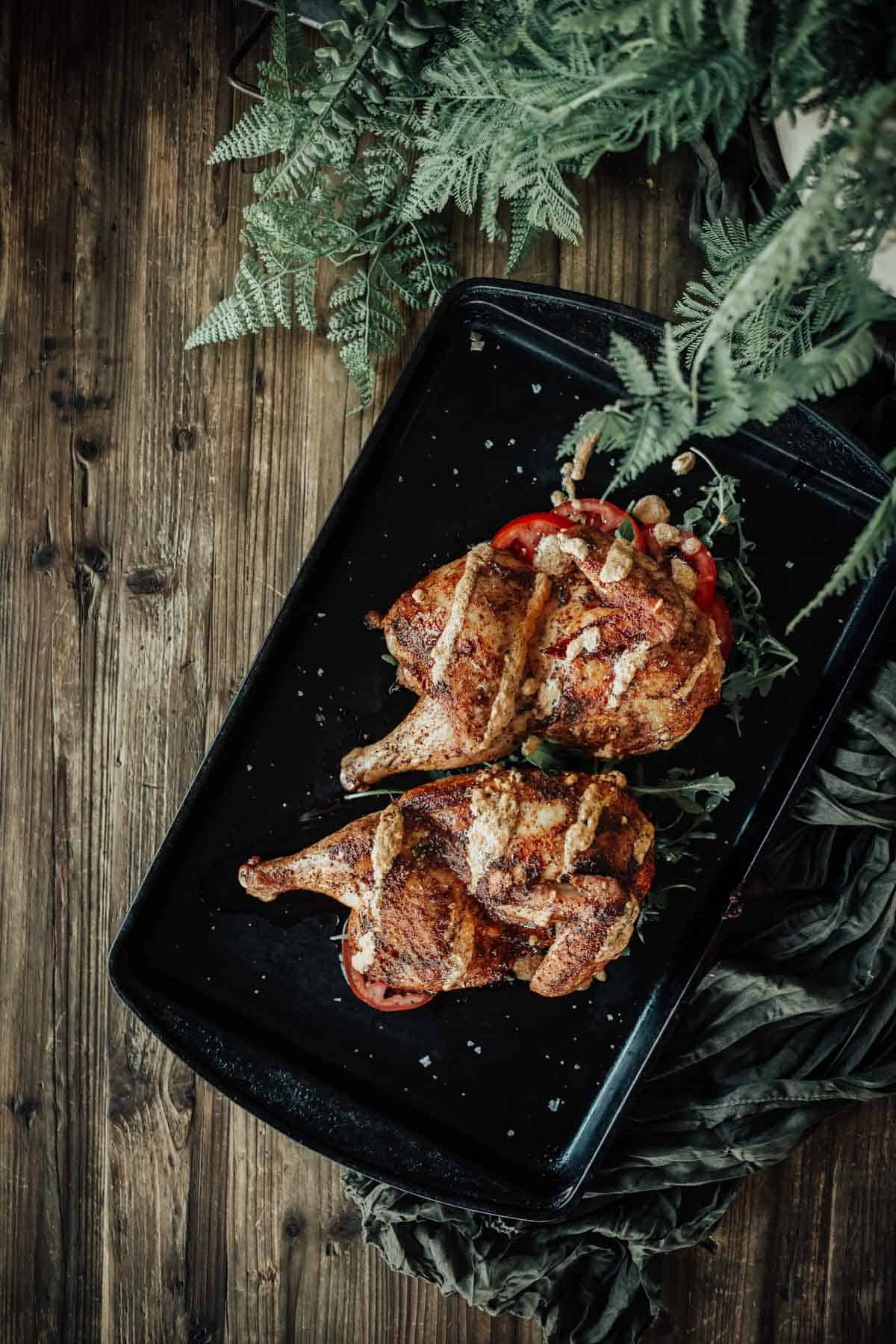 Two grilled chickens stuffed with vegetables are placed side by side on a black tray, with green leafy plants partially visible at the top right corner. The wooden table surface is visible underneath.