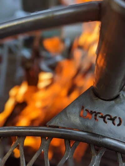 Close-up of a Breeo fire pit with the visible "breeo" logo etched into the metal, set against a background of dancing flames—perfect for enhancing your camp kitchen experience.