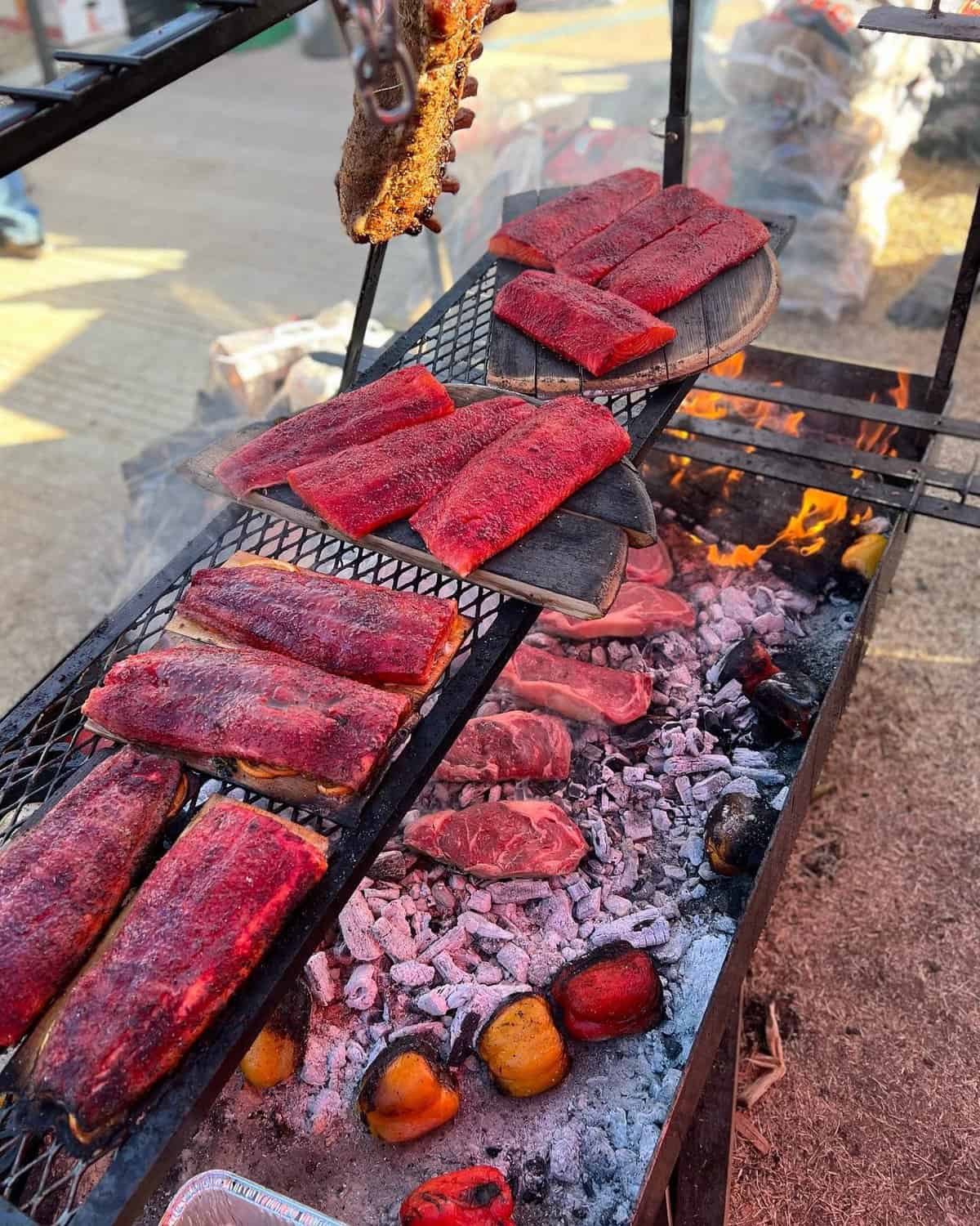 Various cuts of meat are being grilled over an open flame with peppers visible below on the charcoal.