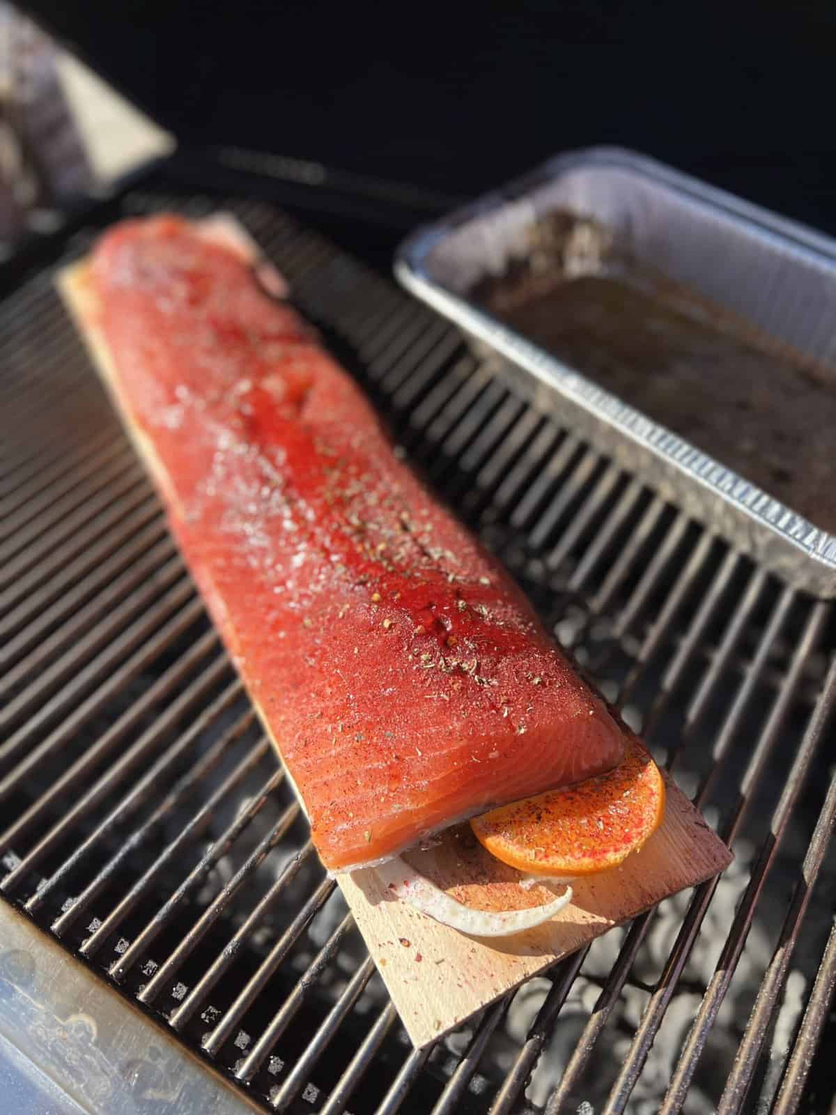 A large fillet of seasoned salmon on a cedar plank, considered the best wood for smoking salmon, is topped with an orange slice and cooking on a grill next to a foil pan.