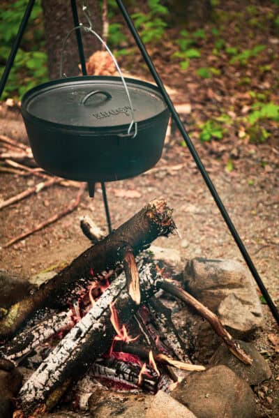 A black cast iron pot hangs over a campfire on a tripod, creating the heart of a rustic camp kitchen. Burning wood and embers glow below, while trees and lush foliage paint the background.