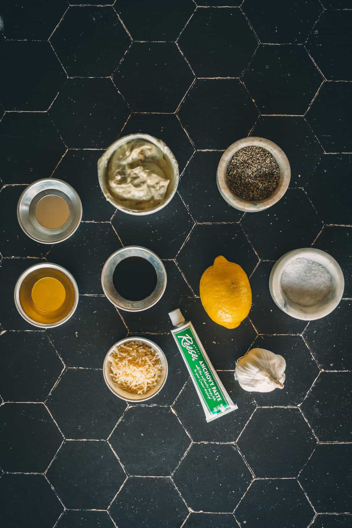 An assortment of ingredients for this dressing recipe including a lemon, garlic, a tube of anchovy paste, grated cheese, mayonnaise, black pepper, and various sauces arranged on a black tiled surface.