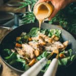 A hand pouring creamy dressing over a Caesar salad with croutons, grilled chicken, Parmesan cheese, and romaine lettuce, on a dark tabletop with greenery in the background.