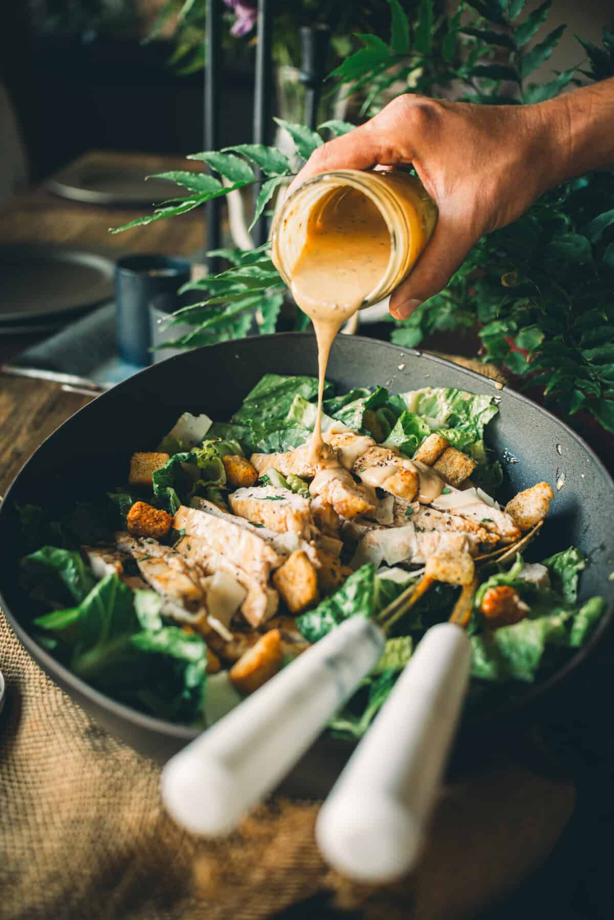 A hand pouring creamy dressing over a Caesar salad with croutons, grilled chicken, Parmesan cheese, and romaine lettuce, on a dark tabletop with greenery in the background.
