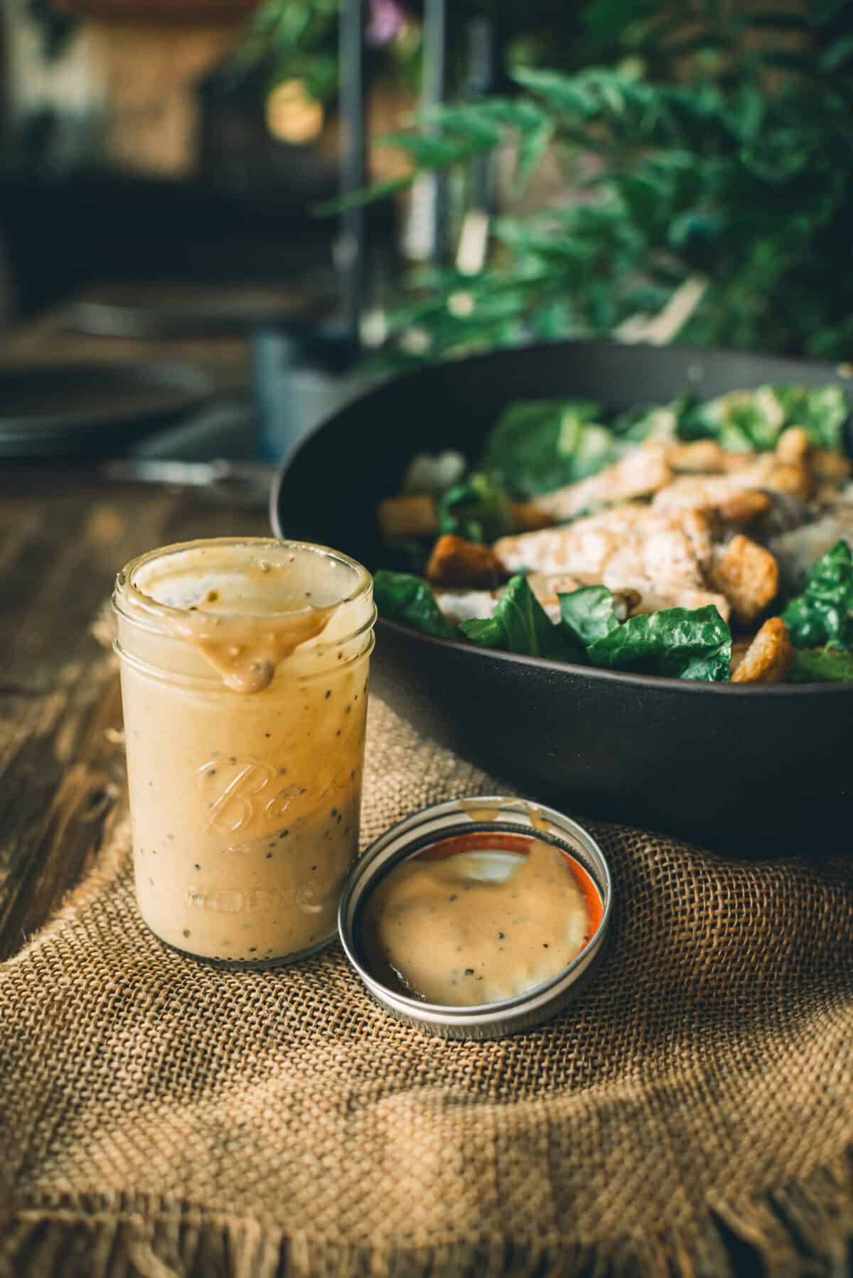 A jar of creamy dressing with the lid removed sits on a table next to a plate containing salad and chicken.