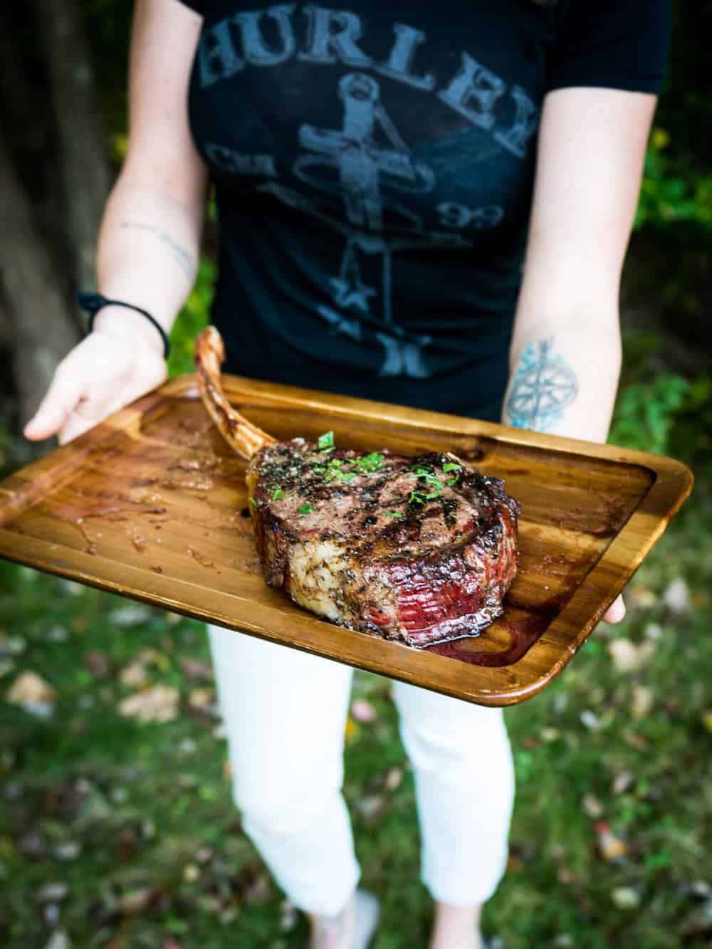 A person wearing a black t-shirt and white pants holds a wooden tray with a grilled tomahawk steak garnished with herbs.