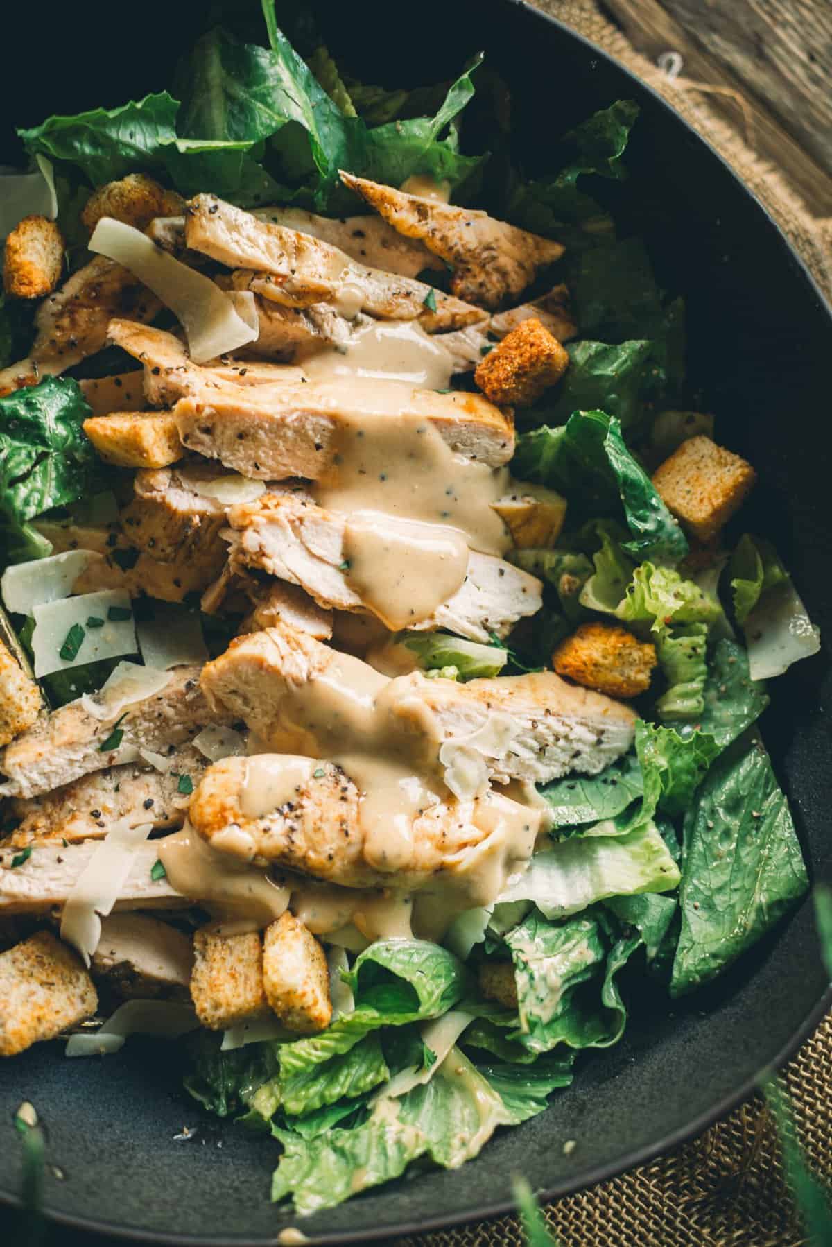 A close up image of a Caesar salad with grilled chicken, croutons, Parmesan cheese, and Caesar dressing in a black bowl.